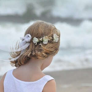 May include: A young girl with blonde hair wearing a white dress and a flower crown with white flowers.