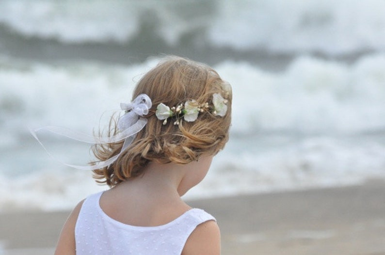 May include: A young girl with blonde hair wearing a white dress and a flower crown with white flowers.