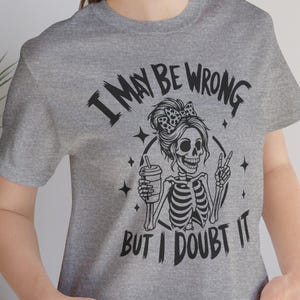 May include: A heather gray t-shirt featuring a skeleton graphic with a coffee cup and a peace sign. The text on the shirt reads "I MAY BE WRONG BUT I DOUBT IT". The design is in black.