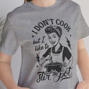 May include: A heather gray t-shirt featuring a retro-style illustration of a woman stirring a pot. The text on the shirt reads "I don't cook but I like to stir the pot."