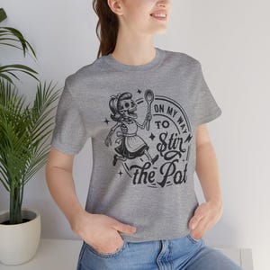Puede incluir: Camiseta gris con una ilustración en blanco y negro de una mujer esqueleto sosteniendo una cuchara, con el texto "On My Way To Stir the Pot". El diseño incluye estrellas y un borde circular.