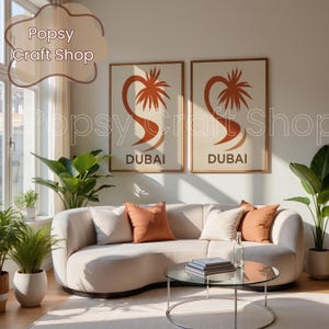 May include: Two framed art prints with a beige background, featuring a stylized palm tree design in orange and the word "DUBAI" in a bold font. The prints hang above a curved beige sofa with orange and cream pillows.