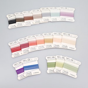 May include: A selection of silk thread spools in diverse shades, such as black, brown, and red. Each spool is marked with "100% Silk" and a needle. The spools are displayed on a light grey surface, perfect for stringing pearls and beads.