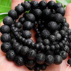 May include: A close-up of a strand of black lava rock beads. The beads are round with a porous texture, varying in size. The beads are strung together, creating a necklace or bracelet. The background is a hand holding the beads.