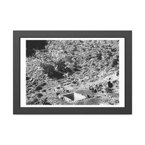 Puede incluir: Impresión enmarcada en blanco y negro de un paisaje con una estructura de piedra y figuras. La imagen captura una ladera con vegetación escasa y un pequeño edificio. Varias figuras son visibles, lo que sugiere un tema histórico o documental.