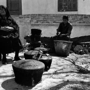 Pode incluir: Uma fotografia a preto e branco mostrando uma pessoa com roupa escura sentada numa cadeira, enquanto outra pessoa trabalha com vários recipientes e potes de metal grandes, redondos e escuros dispostos no chão num ambiente exterior.