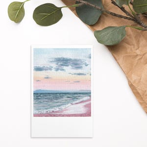May include: A rectangular art print featuring a watercolor painting of a coastal scene at sunset. The image displays a pink sandy beach, turquoise ocean waves, and a soft sky with pastel pink, orange, and blue hues under scattered dark clouds.