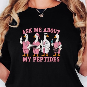 May include: A black t-shirt featuring a graphic of four white geese dressed in medical attire, including scrubs and a lab coat. The text above and below the geese reads, "ASK ME ABOUT MY PEPTIDES" in pink lettering. The geese are depicted with various medical accessories.