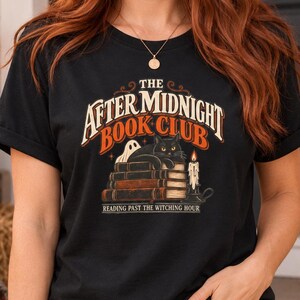 Könnte beinhalten: Ein schwarzes Kurzarm-T-Shirt mit einem Grafikdruck, der den Text THE AFTER MIDNIGHT BOOK CLUB und READING PAST THE WITCHING HOUR zeigt. Das Design umfasst eine schwarze Katze auf einem Bücherstapel, ein kleines Gespenst und eine brennende Kerze.
