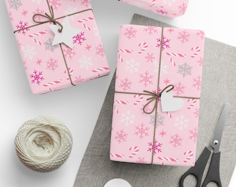 May include: Pink wrapped presents with snowflake and candy cane patterns, tied with brown twine and white heart-shaped tags. A ball of twine, scissors, and blank tags are also present, suggesting a festive theme.