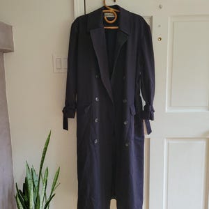 May include: A long, dark blue trench coat with a double-breasted closure, a belt, and two pockets. The coat has a classic design and is made of a smooth, high-quality fabric.