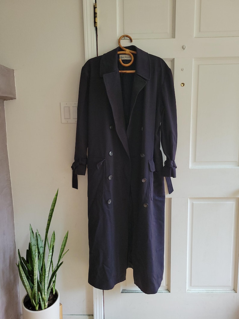 May include: A long, dark blue trench coat with a double-breasted closure, a belt, and two pockets. The coat has a classic design and is made of a smooth, high-quality fabric.