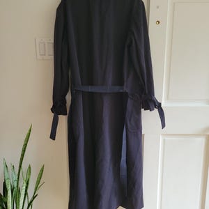 May include: A long, dark blue trench coat with a belt and a tie at the cuffs. The coat is hanging on a wooden hanger.