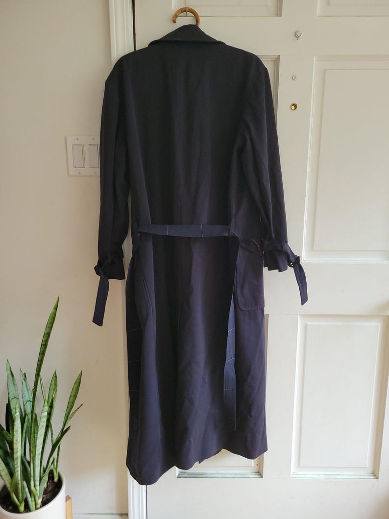May include: A long, dark blue trench coat with a belt and a tie at the cuffs. The coat is hanging on a wooden hanger.