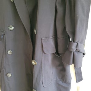 May include: A long, black double-breasted coat with a single pocket on the left side. The coat has a belt and the sleeves have a unique cuff detail.