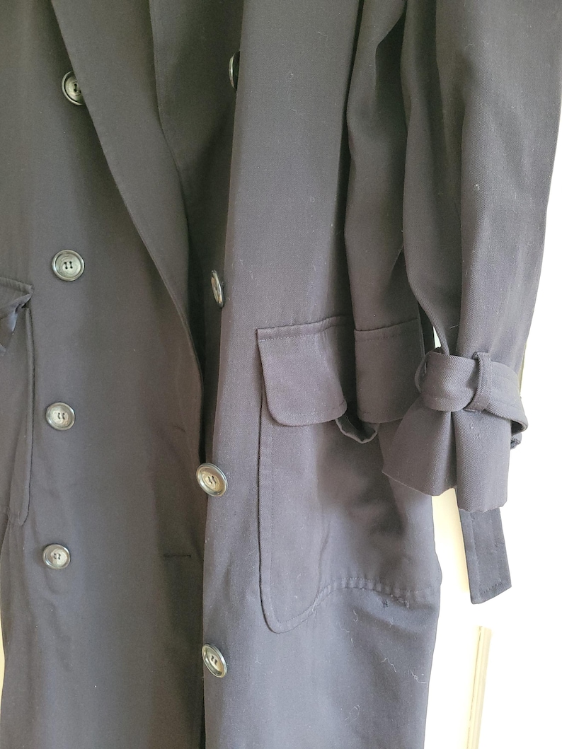 May include: A long, black double-breasted coat with a single pocket on the left side. The coat has a belt and the sleeves have a unique cuff detail.
