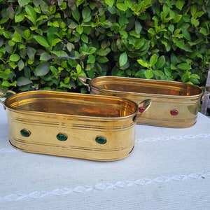May include: Two oval brass planters with green and red gemstone accents. The planters have a hammered texture and are sitting on a white tablecloth.
