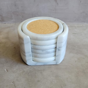 Puede incluir: Un juego de seis posavasos de mármol blanco con un centro de corcho natural, apilados en un soporte de mármol a juego. Los posavasos son redondos y el soporte tiene una base cuadrada. El mármol tiene vetas grises.