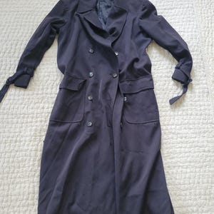 May include: A long black double-breasted coat with long sleeves and pockets. The coat has a tie at the wrists and a label that reads "Anne Klein II" on the collar.