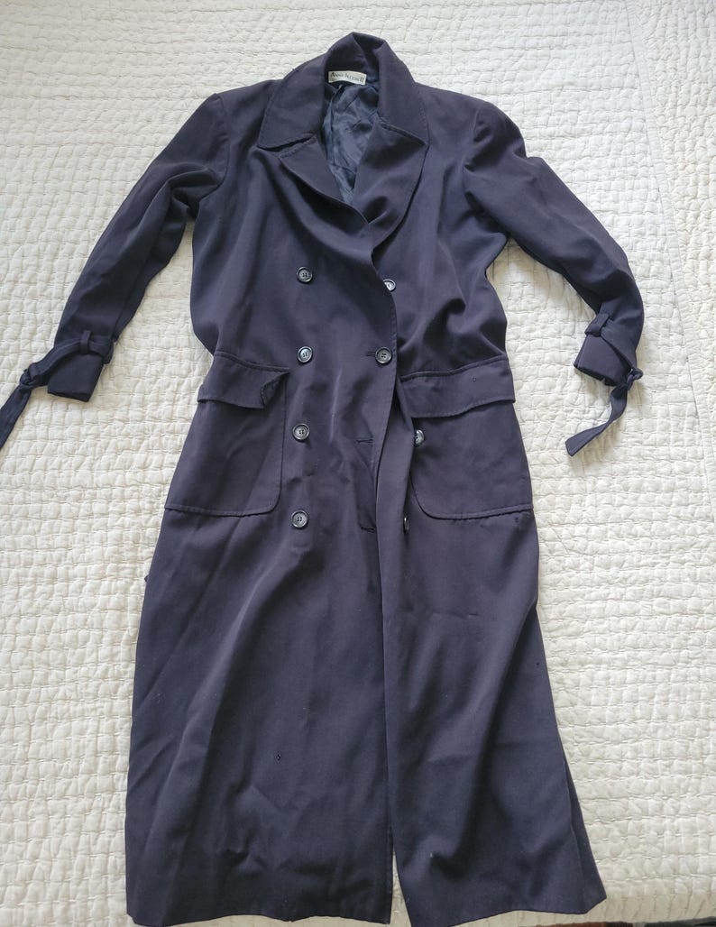 May include: A long black double-breasted coat with long sleeves and pockets. The coat has a tie at the wrists and a label that reads "Anne Klein II" on the collar.