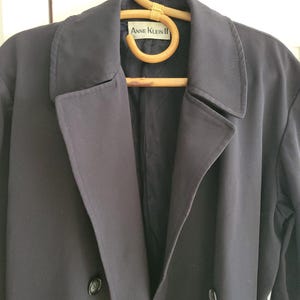 May include: A black double-breasted coat with a label that reads "Anne Klein II" hanging on a wooden hanger.