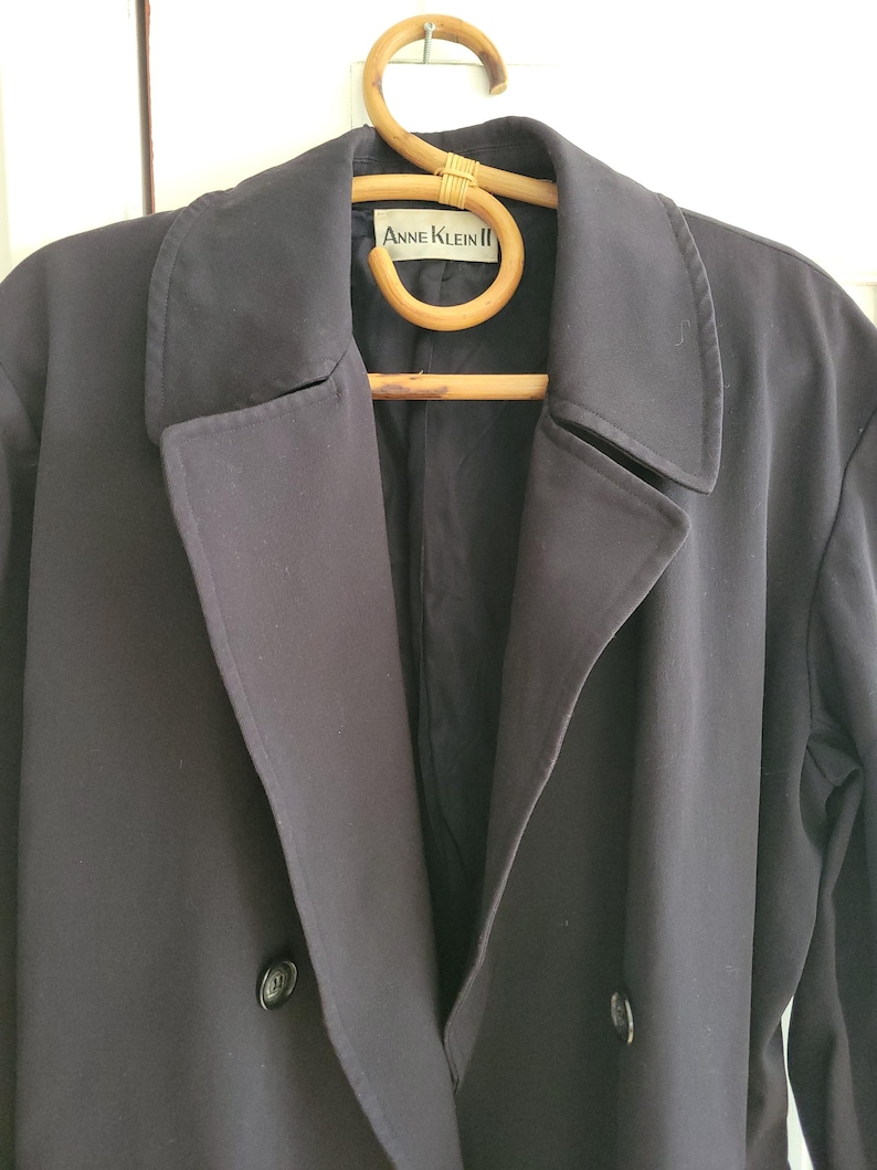May include: A black double-breasted coat with a label that reads "Anne Klein II" hanging on a wooden hanger.