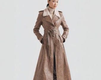May include: A long, brown leather trench coat with a belted waist and a faux fur collar. The coat has a double-breasted design with button closures and pockets. The model is wearing a turtleneck sweater underneath.