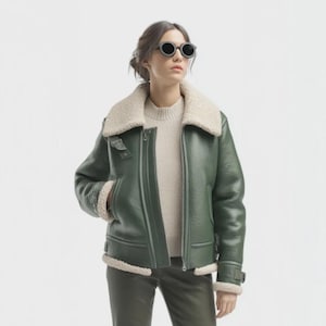 Peut inclure: Veste en cuir vert avec un col et une doublure en peau de mouton crème. La veste a une fermeture éclair, des poches latérales et des détails de boucles sur les manches. La personne porte des lunettes de soleil sombres.