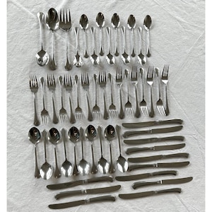 May include: A large collection of stainless steel flatware including various sizes of spoons, forks, and knives. The set features polished metal surfaces with simple, smooth handles arranged in rows on a white fabric surface.