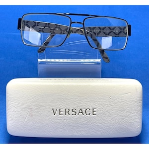 May include: Black framed spectacles with clear lenses, displayed on a clear stand. The arms have a repeating pattern. A white leather case with the brand name "VERSACE" is below the glasses.