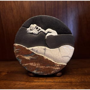 May include: A round ceramic art piece with a landscape design. The top half is black with a white sculpted figure and cloud. The bottom half features brown, white, and blue textured layers. The piece is mounted on small wooden feet.
