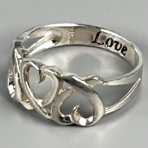 May include: A silver-toned ring featuring an openwork design of two interconnected heart shapes. The interior band of the ring is engraved with the word "Love" in a black, stylized script font, visible against the polished metallic surface.