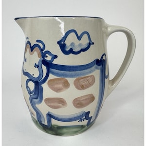 Puede incluir: Jarra de cerámica color crema con un diseño caprichoso de vaca. La vaca está representada con contornos y acentos azules, junto con manchas marrones. La jarra tiene un asa curva y un pico para verter. La base es verde.