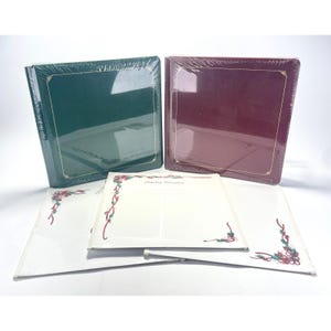 Puede incluir: Dos álbumes de fotos, uno verde oscuro y otro burdeos, están envueltos en plástico. Los álbumes tienen adornos dorados. Se ven páginas sueltas con las palabras "Holiday Memories" y diseños florales.