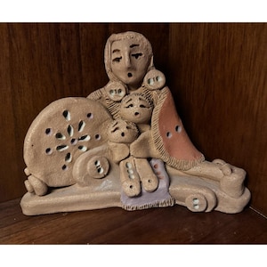 May include: A tan ceramic sculpture depicting a mother and two children. The mother figure has large earrings and a shawl. The sculpture includes a wheel-like object with cut-out designs and a small purple blanket.