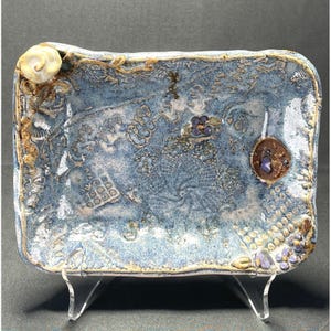 May include: A rectangular ceramic tray with a blue glaze and intricate embossed patterns. The tray features a seashell, floral accents, and a circular brown embellishment with a purple center. The tray is supported by clear acrylic legs.