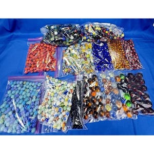 Pode incluir: Uma coleção de bolas de gude em várias cores, incluindo vermelho, azul, verde e amarelo, é exibida em sacos plásticos transparentes. As bolas de gude têm diferentes padrões e tamanhos e estão dispostas em uma superfície azul.