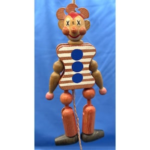 May include: A vintage wooden pull-string toy clown with a painted face, striped torso, and jointed limbs. The clown has a red face, brown and white striped torso with three blue circles, and wooden legs and arms. The toy is suspended by a string.