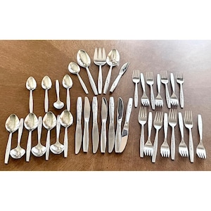 May include: A collection of stainless steel silverware, including spoons, forks, and knives, arranged on a wooden surface. The silverware has a simple, elegant design, with a floral pattern on the handles. The set includes various sizes of spoons and forks.