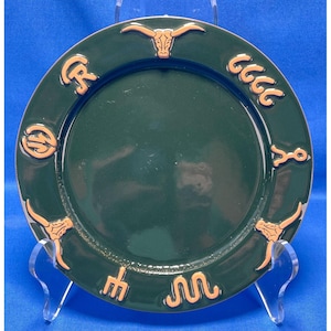 Puede incluir: Un plato redondo de cerámica verde oscuro con un diseño en relieve de color claro a lo largo del borde. El diseño incluye dos cráneos de buey, una horca, una línea ondulada similar a una serpiente y varios símbolos de marcas de ganado.