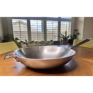 May include: A stainless steel wok pan with a long handle and a small loop handle. The pan is on a wooden table. The wok is round and has a curved shape. The pan's diameter is approximately 30 cm.