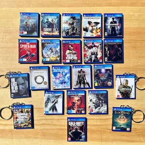 Könnte beinhalten: Eine Sammlung von Miniatur-PS4-Spielhüllen, jede mit einem einzigartigen Cover-Design. Zu den Titeln gehören "God of War", "Spider-Man" und "Grand Theft Auto". Die blauen Hüllen sind mit einem silbernen Schlüsselanhänger versehen, auf einer hellbraunen Oberfläche.