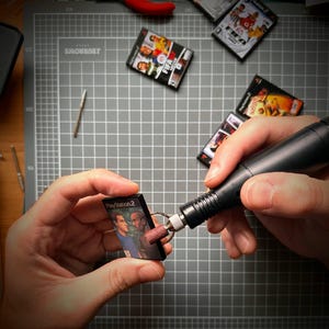 May include: A person is working on miniature PlayStation 2 game cases. The cases, with colourful game covers, are being crafted using a small rotary tool. Several other miniature game cases are scattered on a grey cutting mat.
