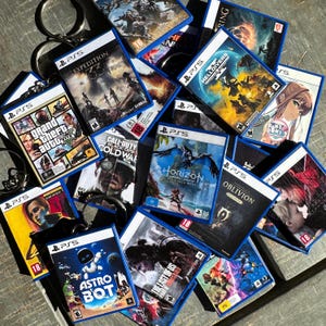 Może przedstawiać: Kolekcja breloków do gier PS5 z różnymi tytułami, takimi jak "Grand Theft Auto", "Horizon Forbidden West" i "Astro Bot". Każdy brelok ma niebieską obwódkę i metalowy pierścień. Gry są oceniane na 16 lub 18.