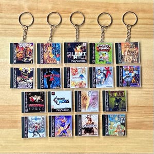May include: A selection of PlayStation game case keyrings. Each keyring showcases a miniature replica of a classic game case, including titles such as "Silent Hill," "Twisted Metal 4," and "Harvest Moon." The keyrings have silver metal rings.
