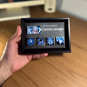 May include: A black rectangular frame containing a rectangular image of Crystal Maiden and four square tiles featuring blue-toned magical ability icons. The text "NTELLIGENCE" and "CRYSTAL MAIDEN" is printed in white on the black background.