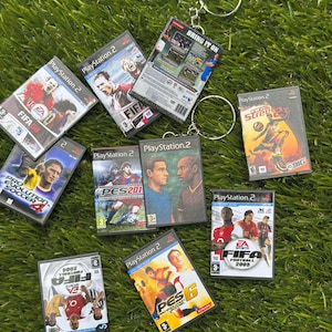 Puede incluir: Miniaturas de juegos de PlayStation 2 con juegos de fútbol como FIFA y PES. Las carcasas, algunas unidas a llaveros, muestran ilustraciones coloridas. Los juegos incluyen FIFA 08 y PES 6. Los artículos están dispuestos sobre una superficie verde.