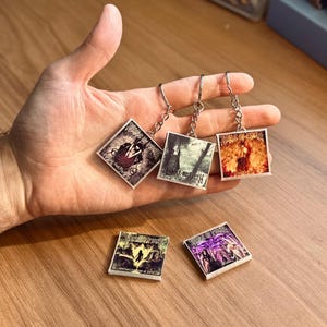 Puede incluir: Llaveros plateados con colgantes cuadrados con arte gótico y texto. Los llaveros tienen una cadena y un anillo. Los colgantes muestran imágenes en tonos rojos, negros, blancos y morados. También se ven dos colgantes cuadrados adicionales.