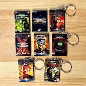 May include: A collection of seven Command & Conquer game case keychains. Each keychain features a different game title, with vibrant cover art and attached to a silver keyring. The keychains are arranged on a light brown wooden surface.