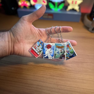Puede incluir: Una mano sosteniendo cinco llaveros con ilustraciones de cajas de juegos en miniatura. Los títulos de los juegos incluyen "Super Mario Bros. U" y "Rayman". Cada llavero tiene un anillo plateado. El fondo incluye luces pixeladas con temática de videojuegos.